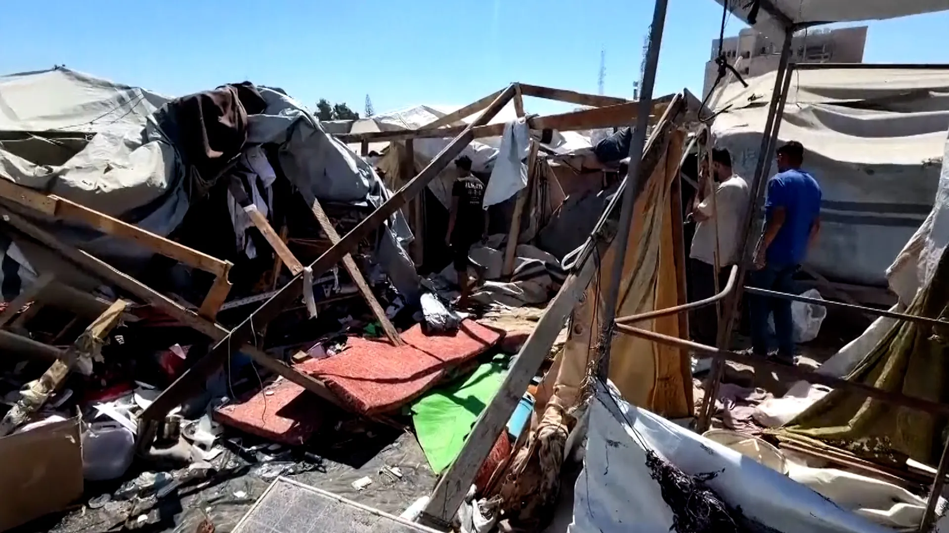 Men inspect a severely damaged displacement camp filled with torn tarps, debris, and broken wooden supports.