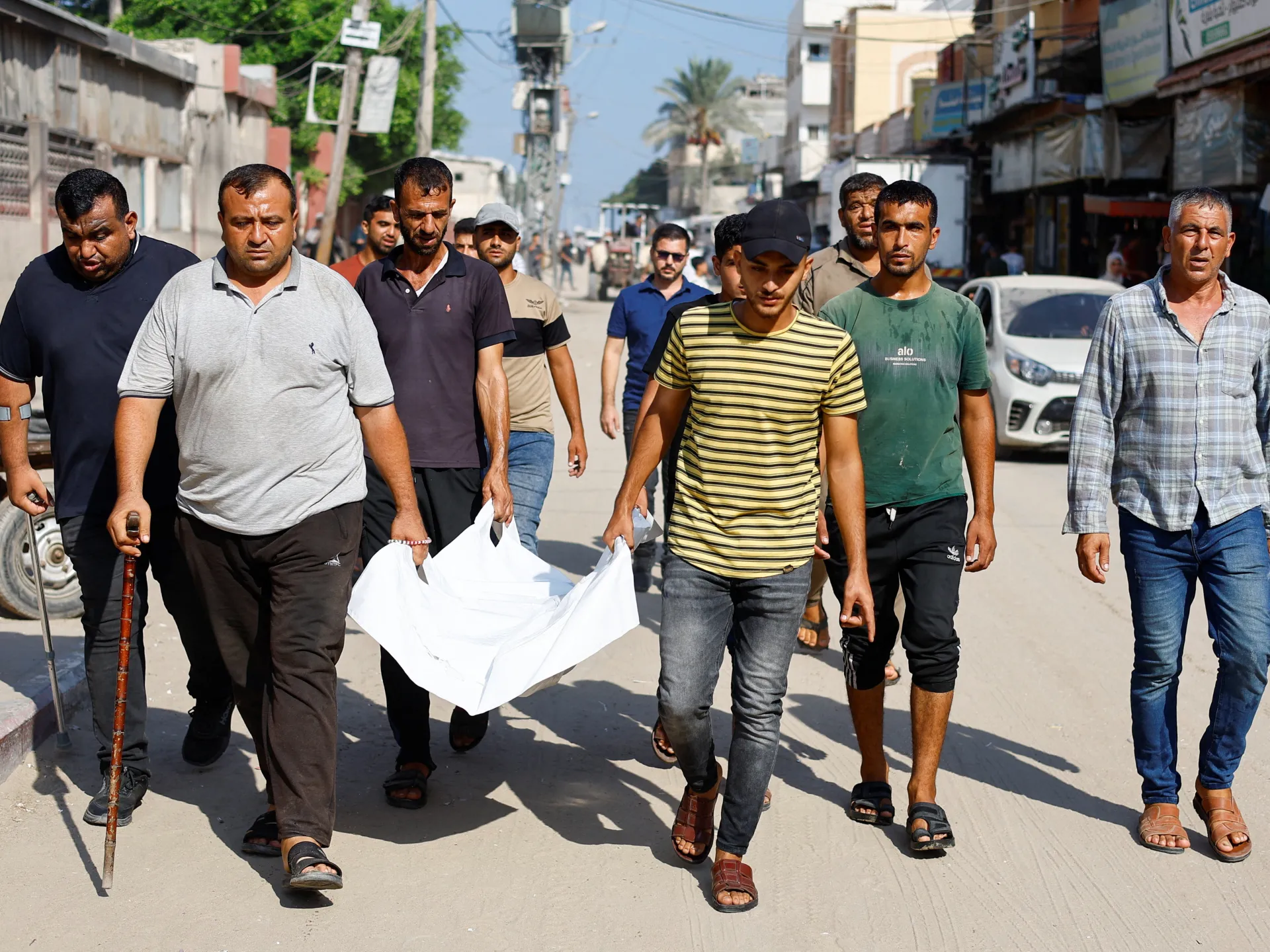 Men carry a white cloth through a sunlit urban street as others walk nearby.