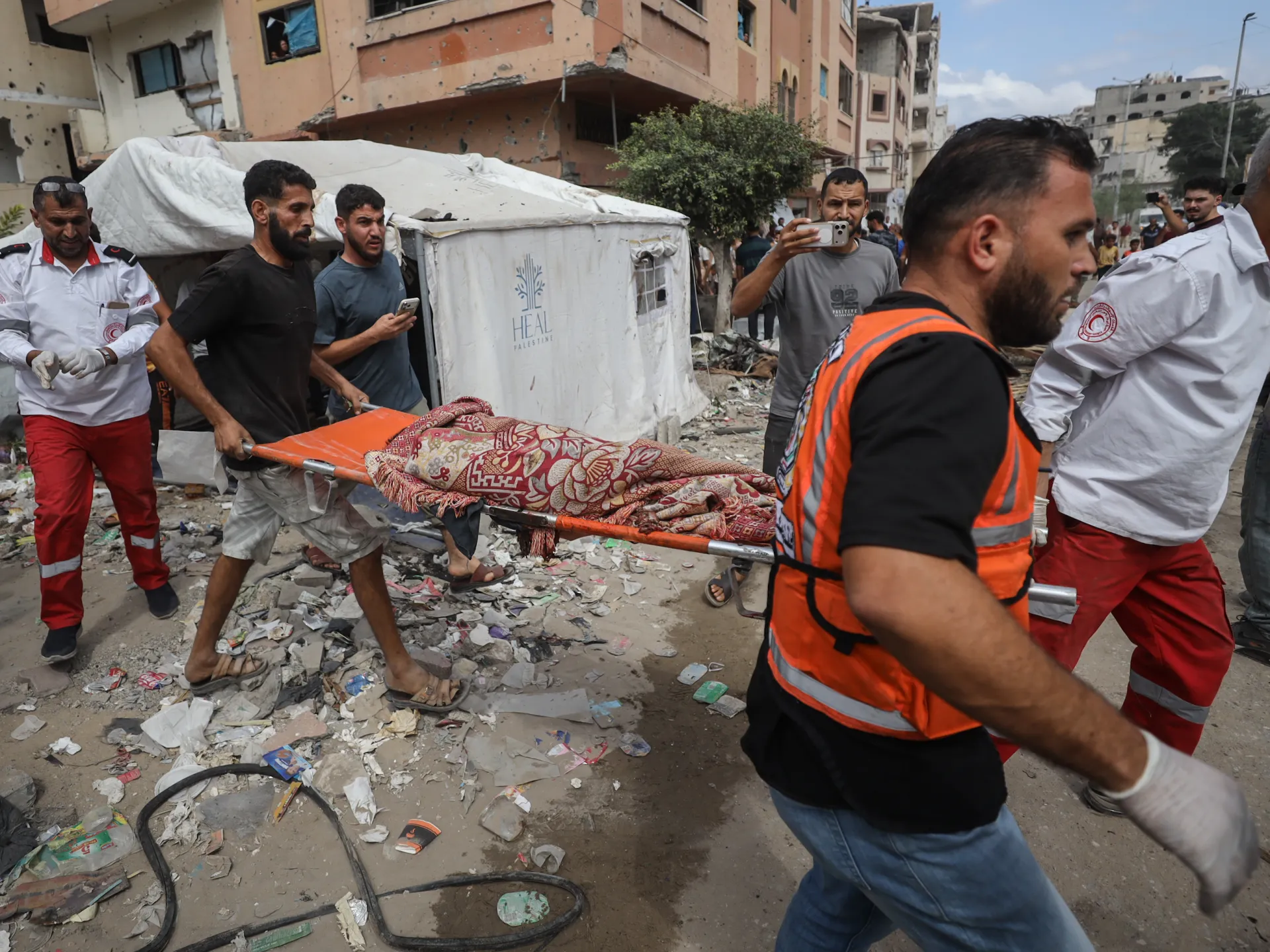 Rescue workers and men carry a blanket-covered stretcher through rubble beside damaged buildings.