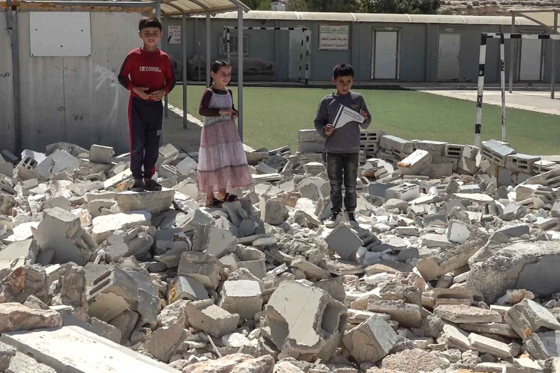 Three children stand on rubble outside a damaged compound, with a sports field behind them.