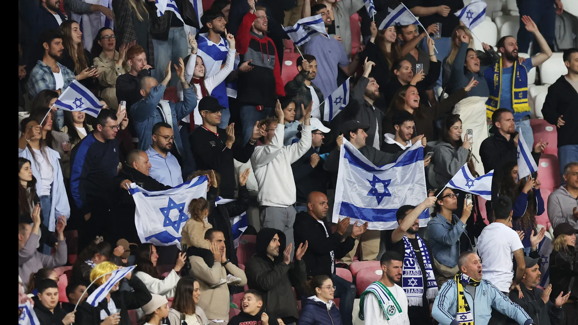 Football fans cheer in stadium seats, waving Israeli flags and wearing team scarves.