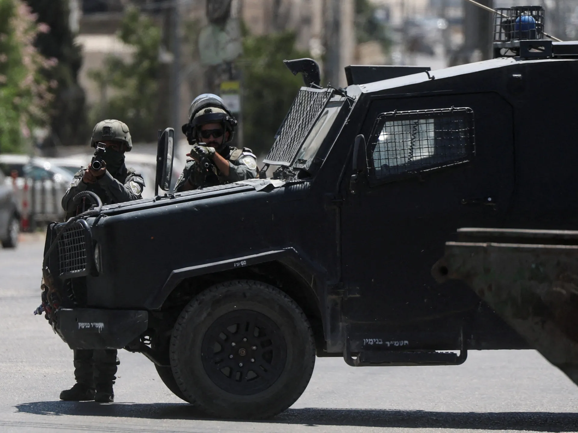 Armed personnel stand beside a black armored vehicle on a sunlit urban street.