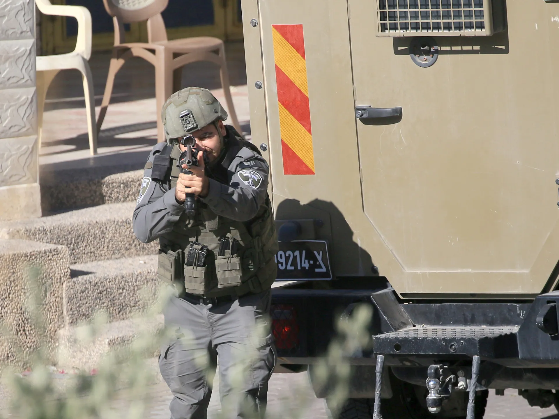 Armed soldier in protective gear aims a rifle beside an armored vehicle.
