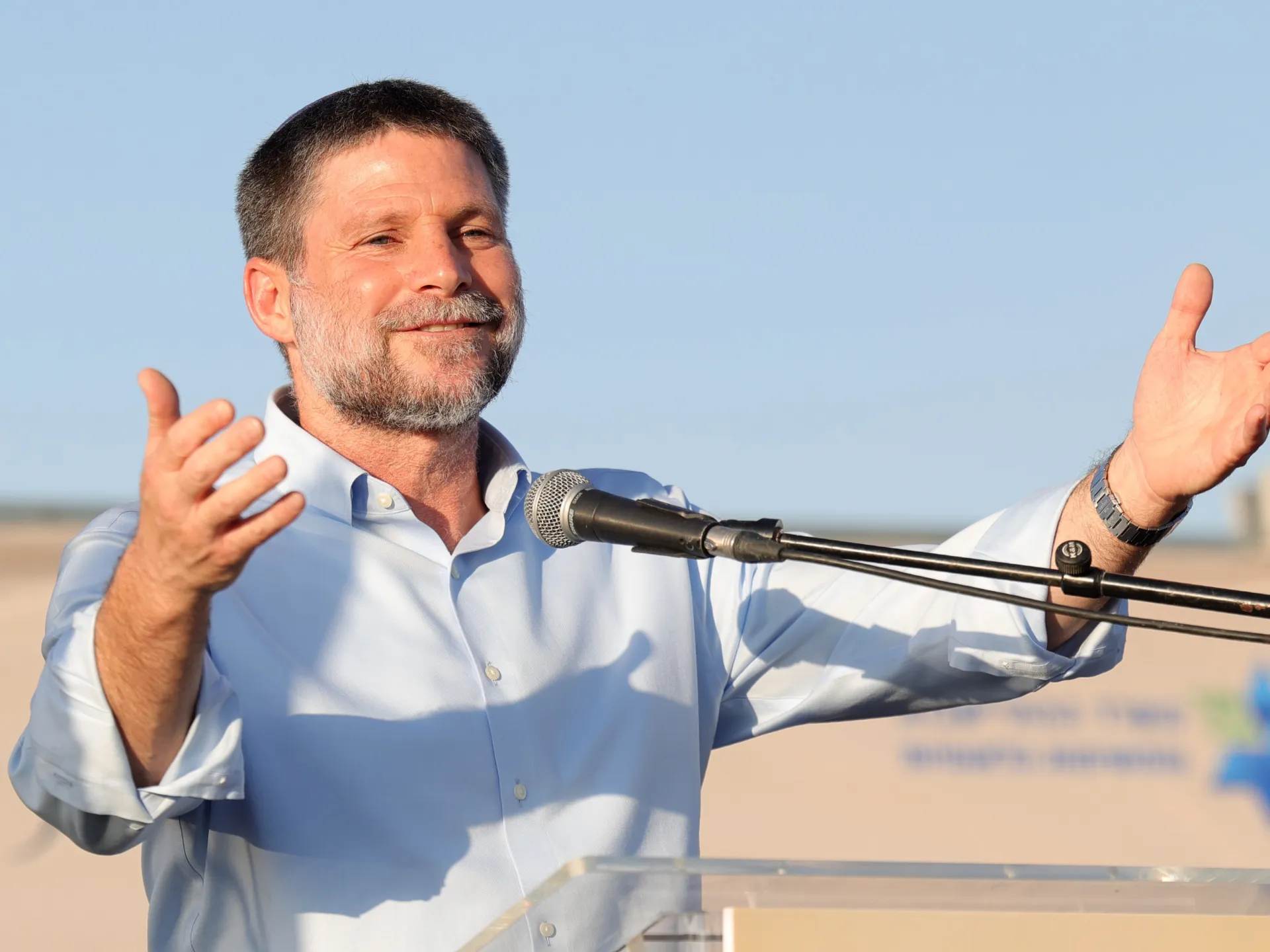 A man in a light blue shirt speaks at a microphone with both hands raised outdoors.