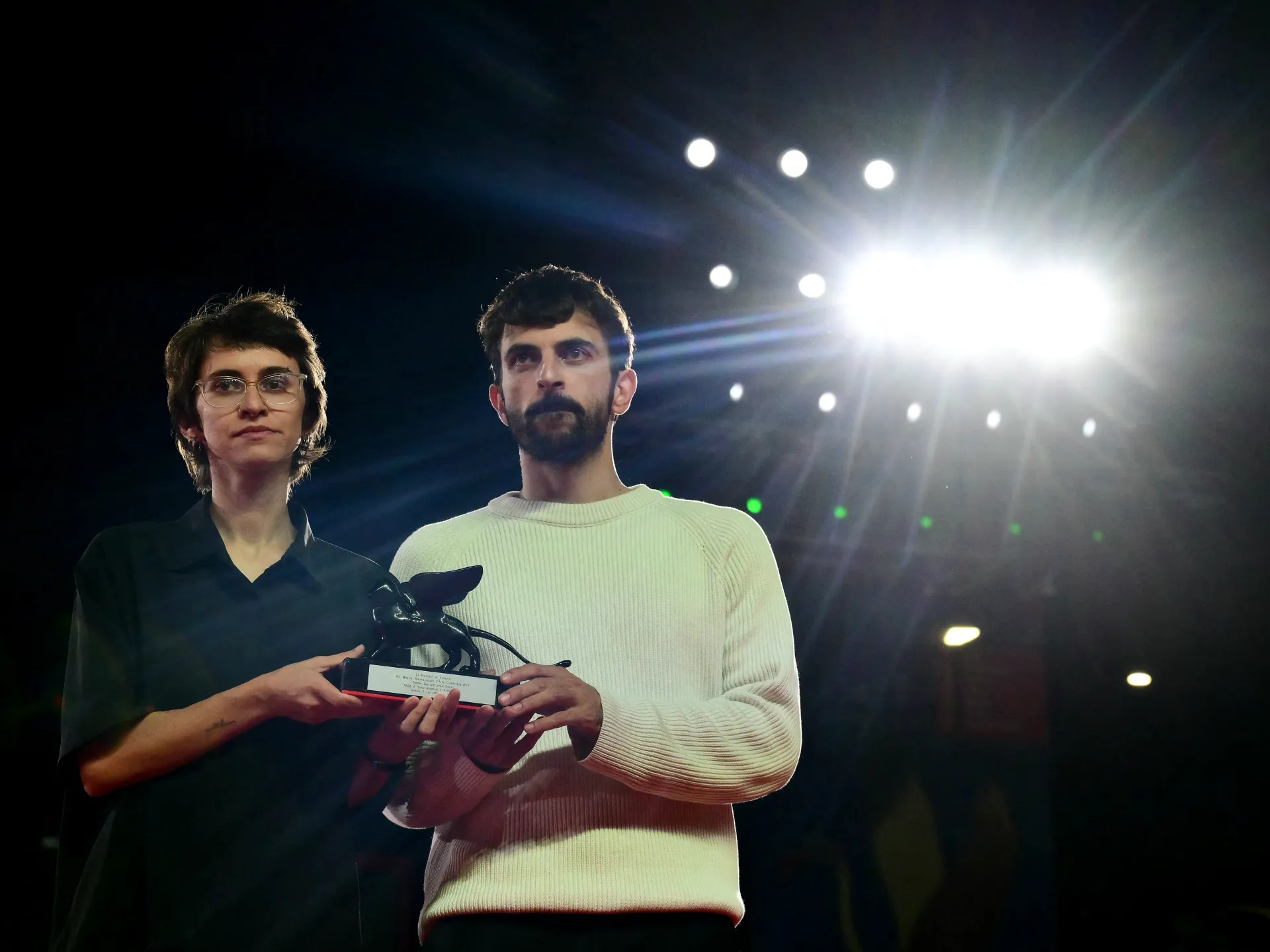 Two people hold a black trophy beneath bright stage lights in a dark venue.