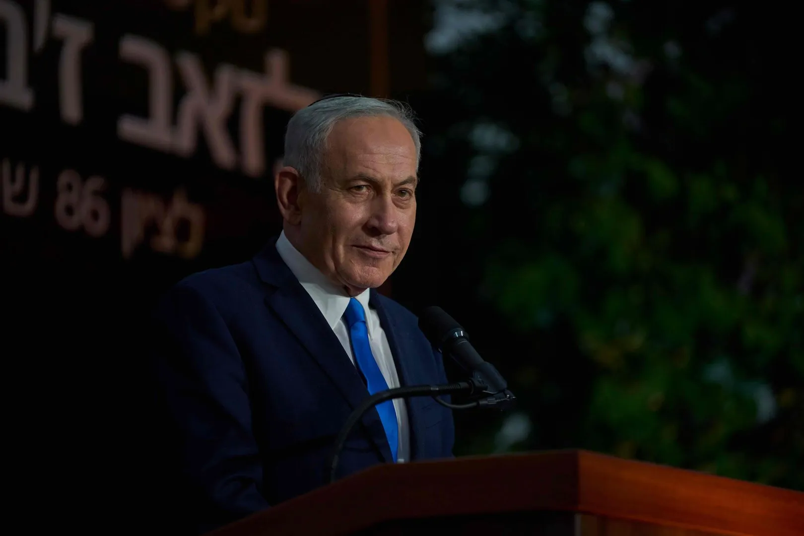 Israeli Prime Minister Benjamin Netanyahu speaks at a podium beside a microphone in a dimly lit setting.