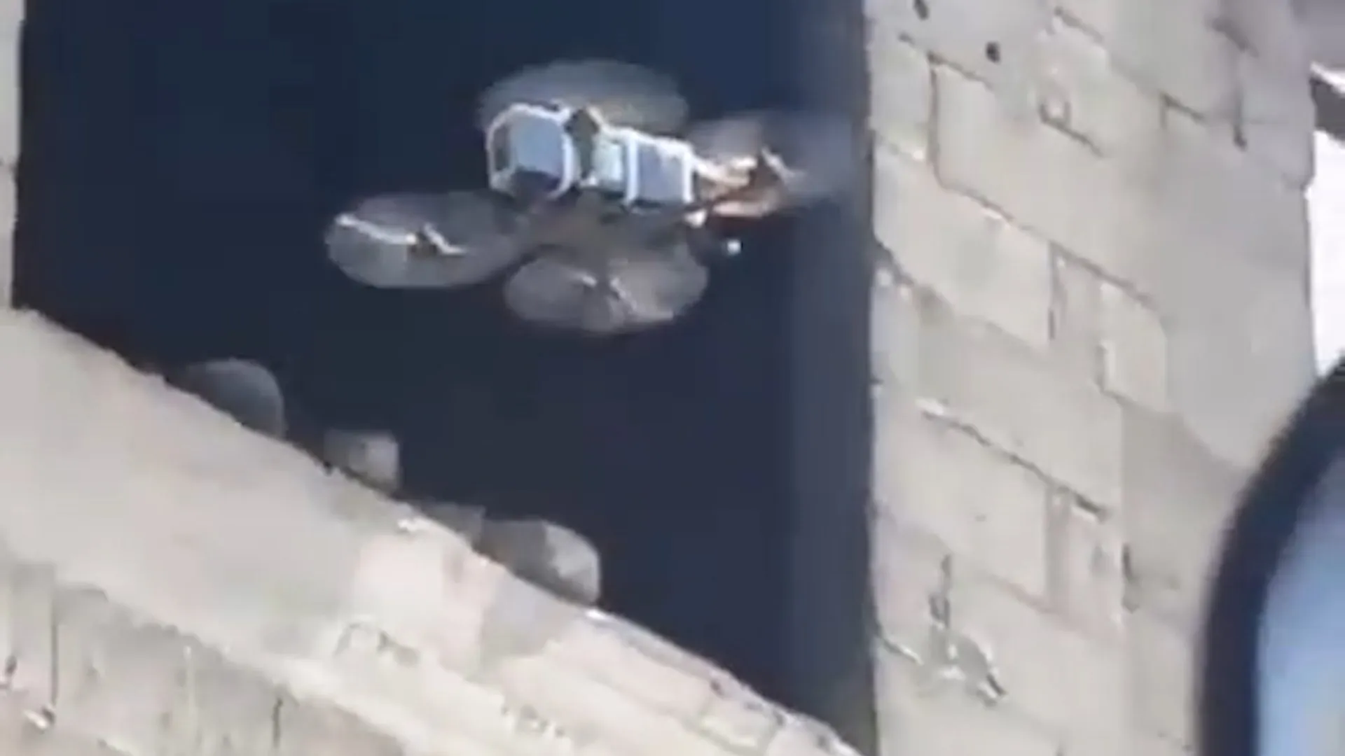 A small quadcopter drone hovers beside a pale brick building above a rooftop.