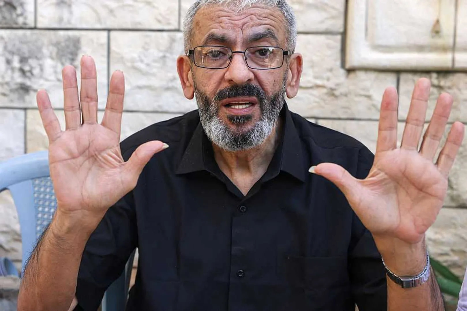 Bearded man wearing glasses and black shirt raises both hands toward the camera.