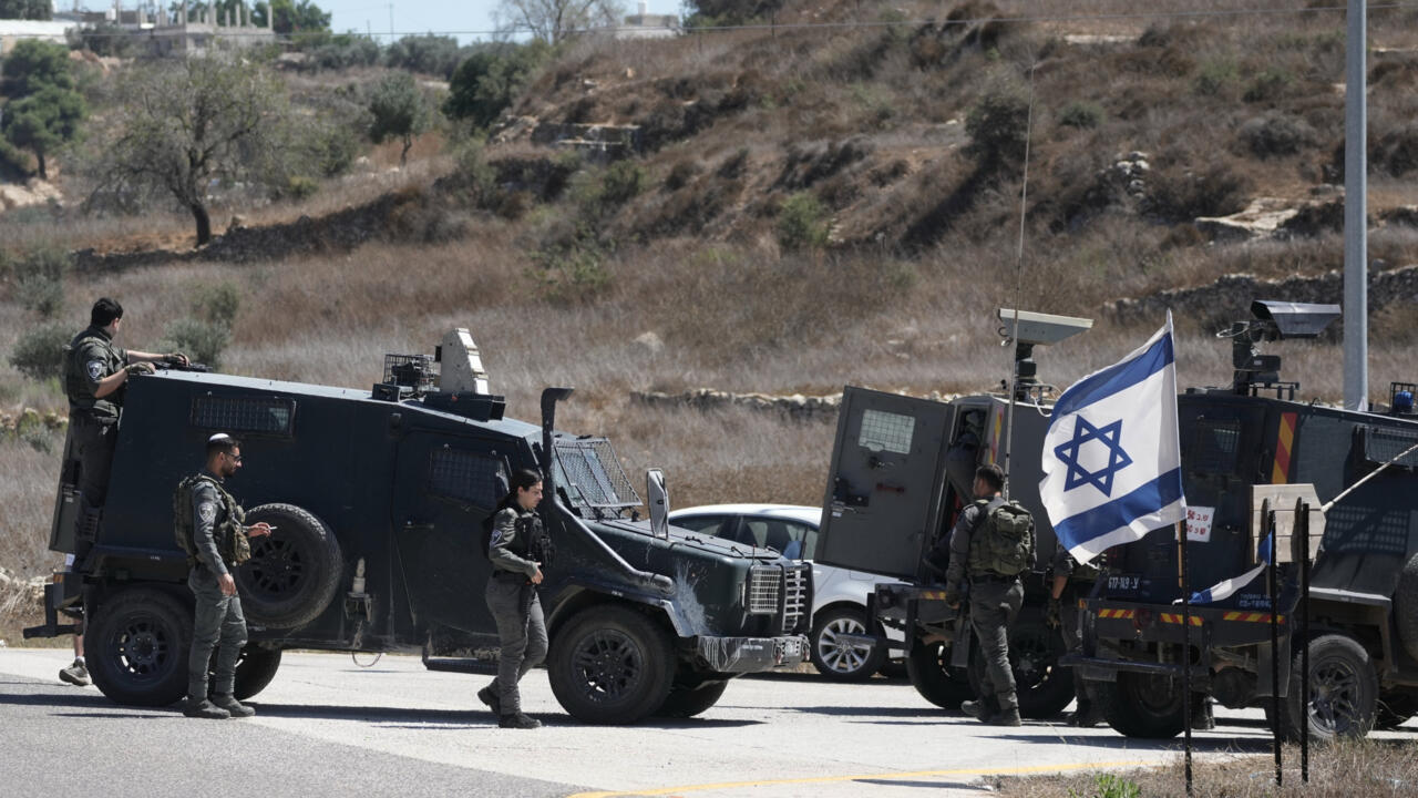 Israeli security personnel stand beside armored vehicles and an Israeli flag on a rural road.