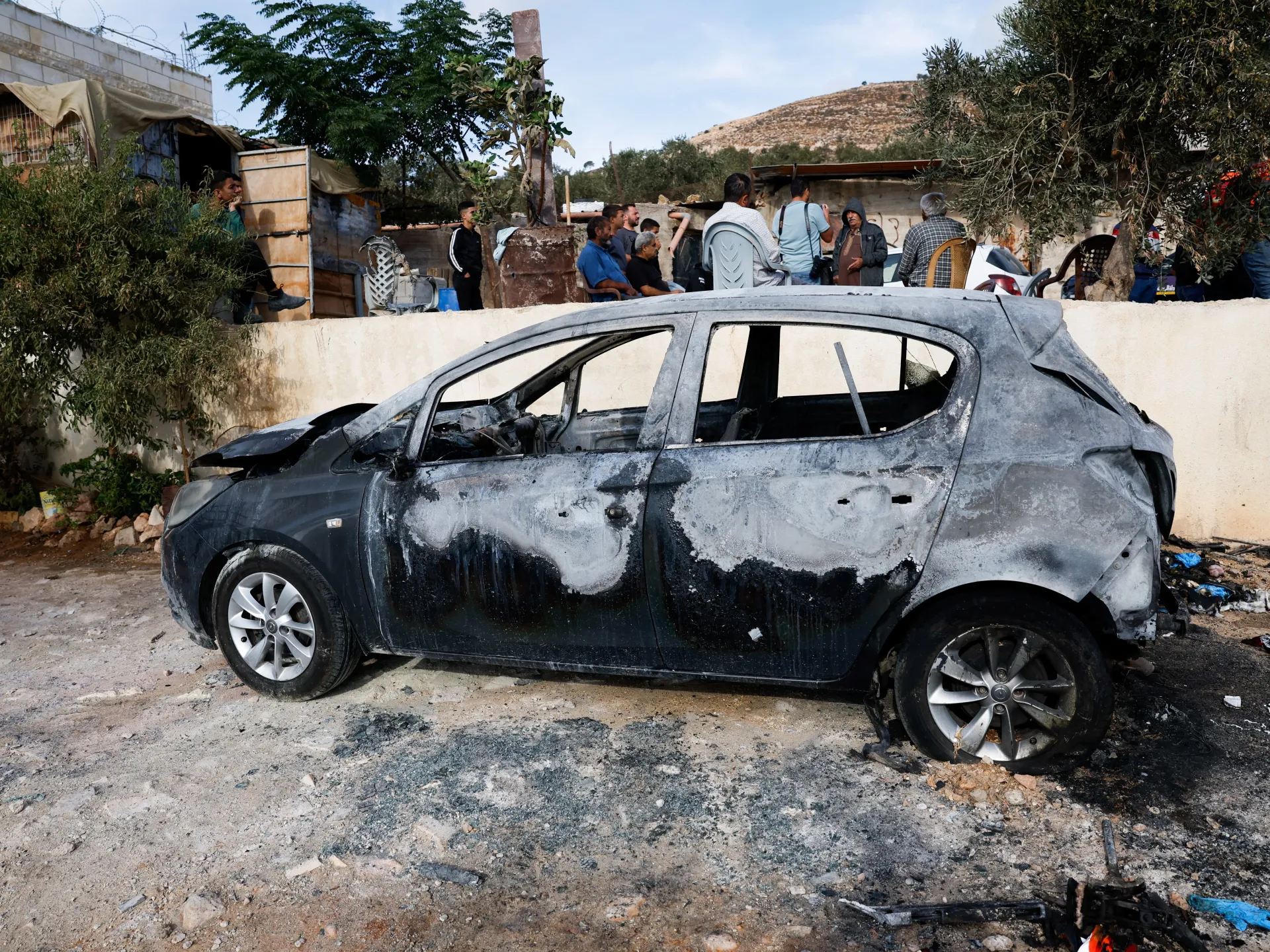 A burned-out car sits in the foreground as several people gather behind a wall.