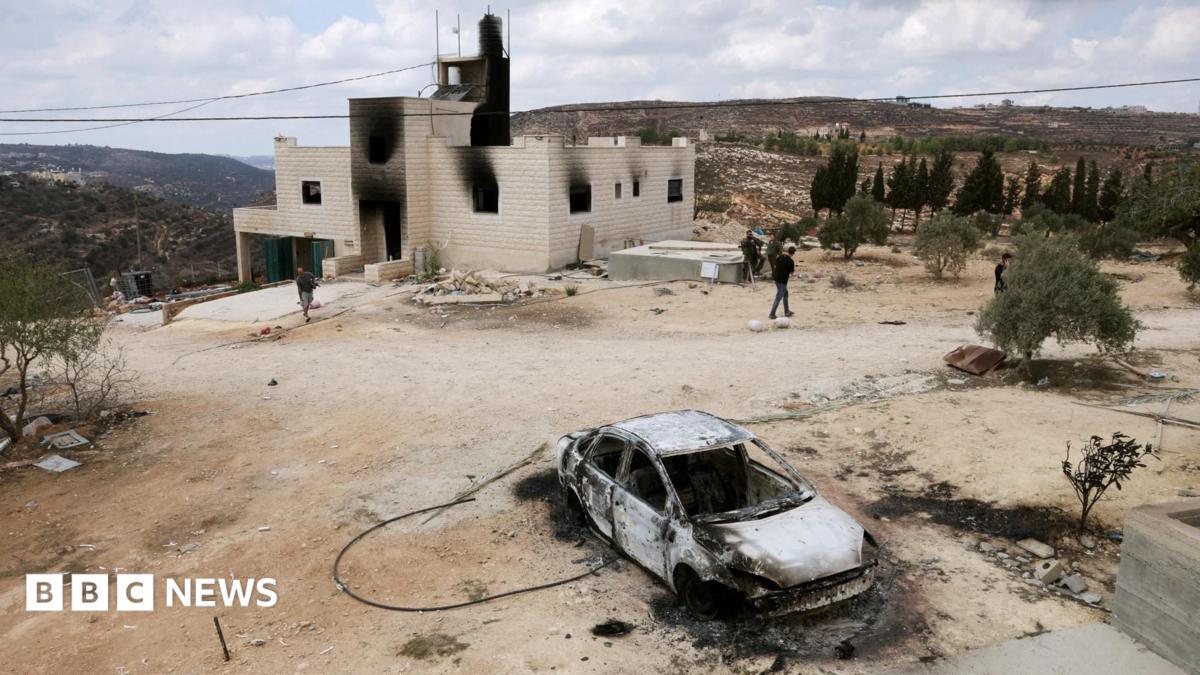 A burned-out car sits in a dirt yard outside a stone house in a hilly area.