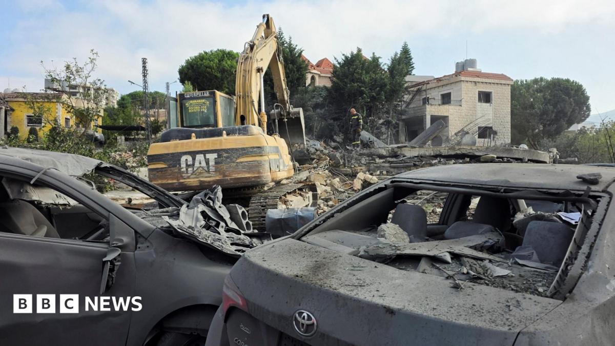 Excavator clears rubble beside destroyed cars and damaged buildings after apparent strikes