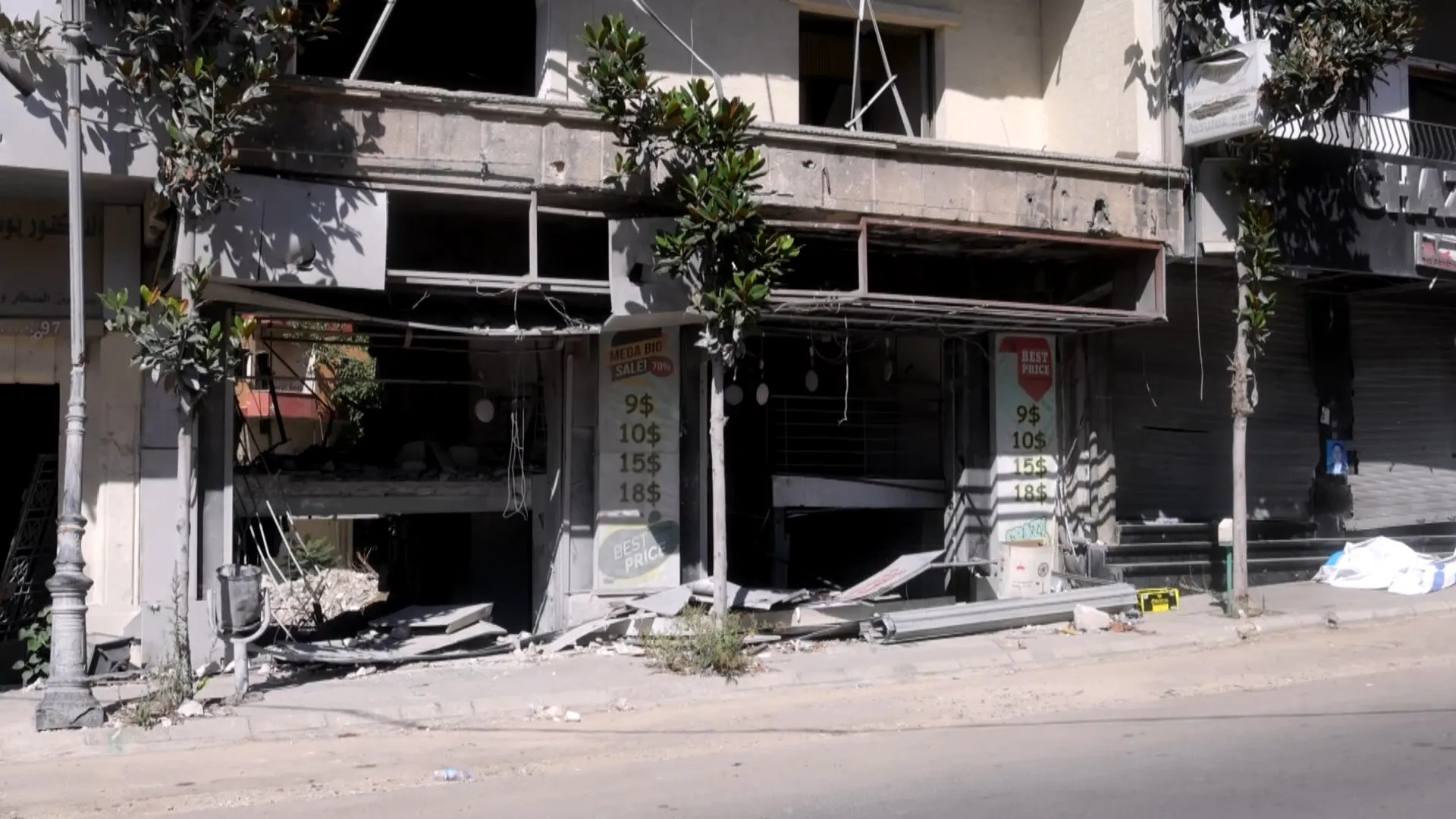 Damaged multi-story storefront with shattered windows, exposed wiring, rubble, and debris along the sidewalk