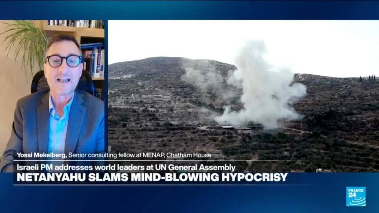 Split-screen television broadcast shows a suited man speaking beside footage of smoke rising over a barren hillside.