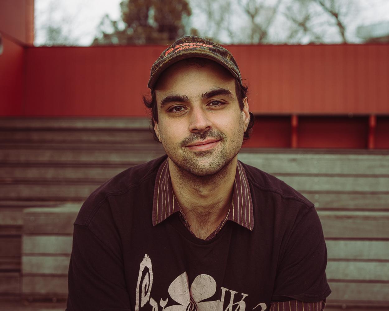 Man wearing a cap and dark shirt sits outdoors in front of red and weathered wooden panels.