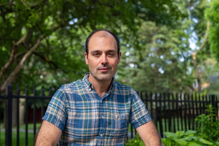 A man in a blue plaid shirt stands outdoors in front of trees and a fence.