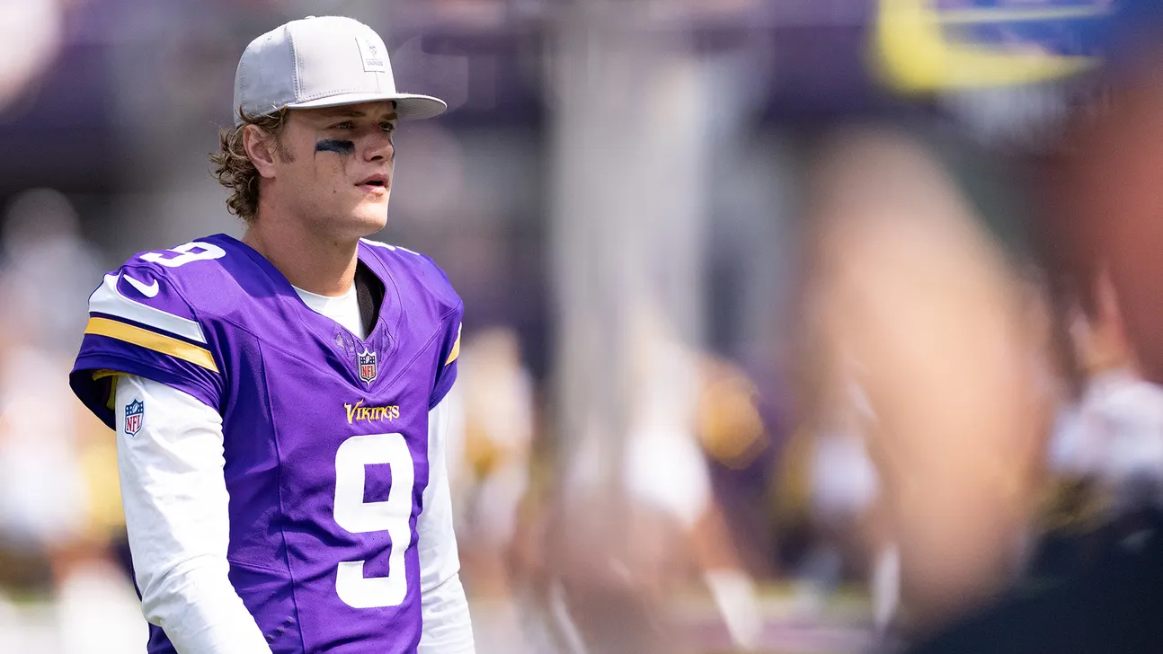 Football player in a purple Vikings jersey and white cap stands on the sideline amid blurred figures.