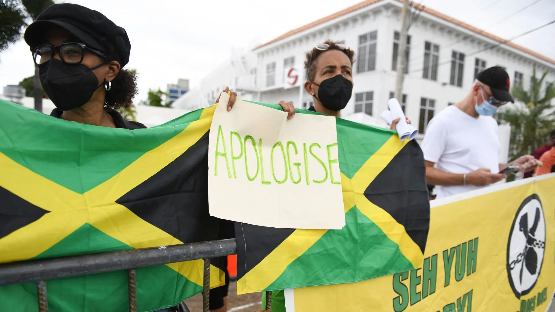 Masked demonstrators hold Jamaican flags and an “APOLOGISE” sign during a public protest.