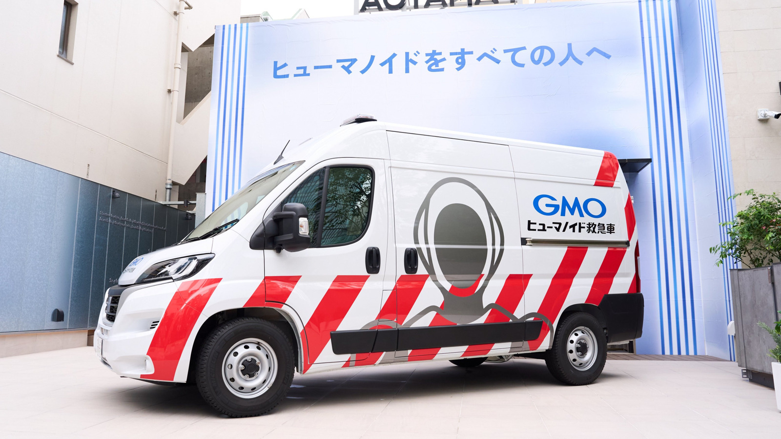 White van with humanoid robot graphic and red stripes displayed before a blue-and-white backdrop