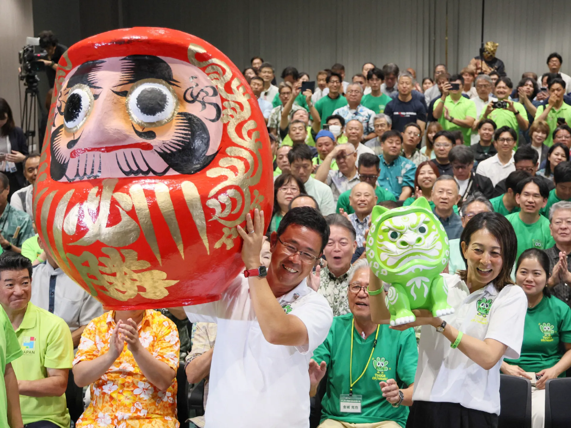Crowd celebrates with large red daruma and green character figures at a public event