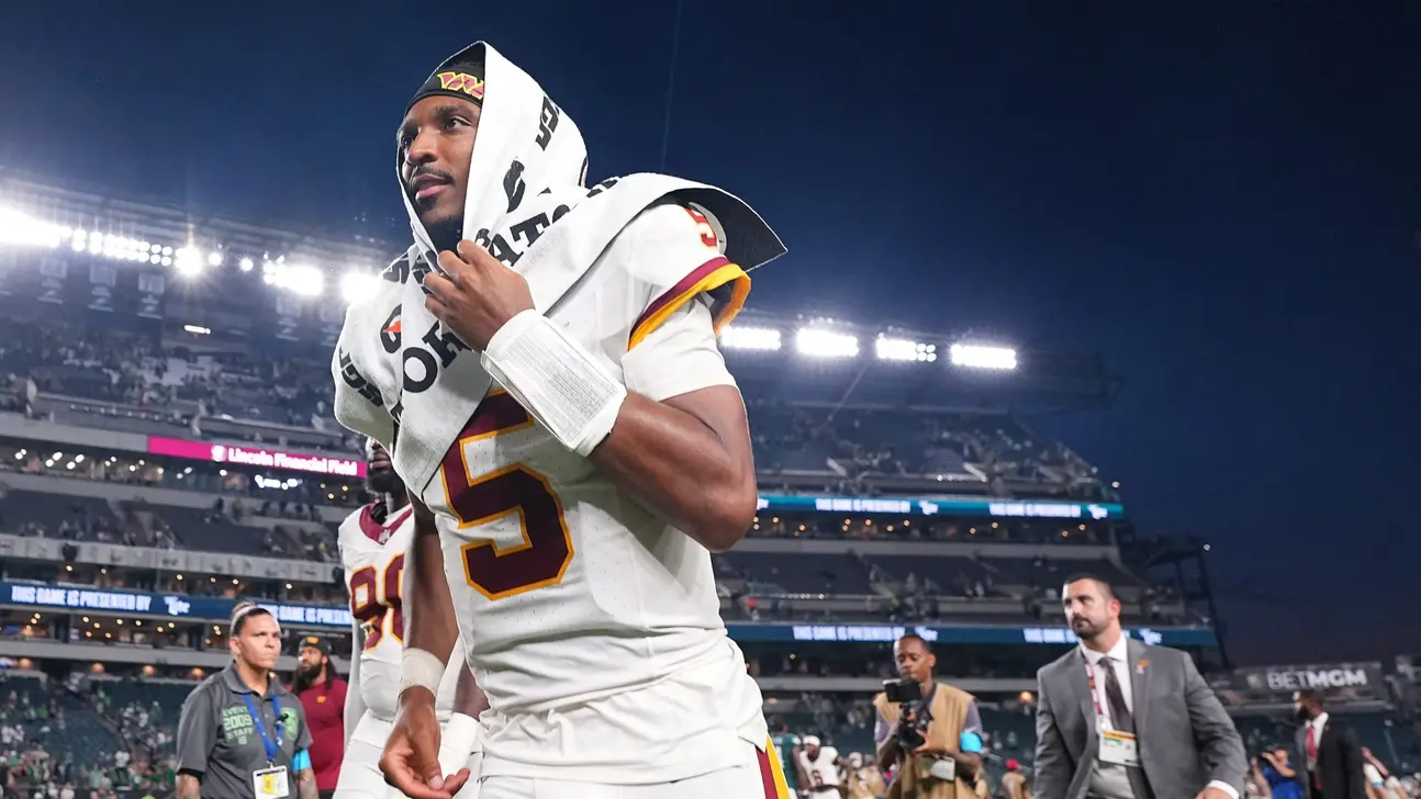 Football player in white uniform walks through stadium wearing a towel over his head.