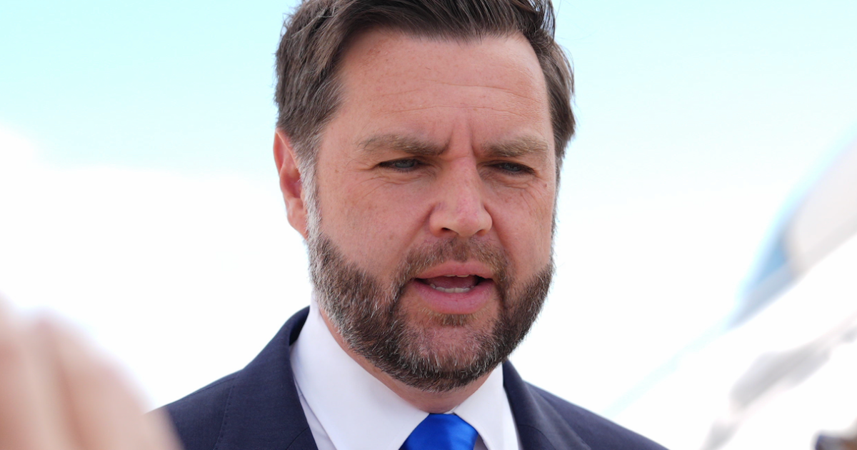 Close-up of a bearded man in a dark suit and blue tie speaking outdoors