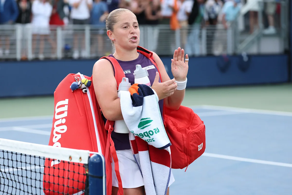 Tennis player gestures while carrying racquets, water bottles, towels, and bags beside the net.