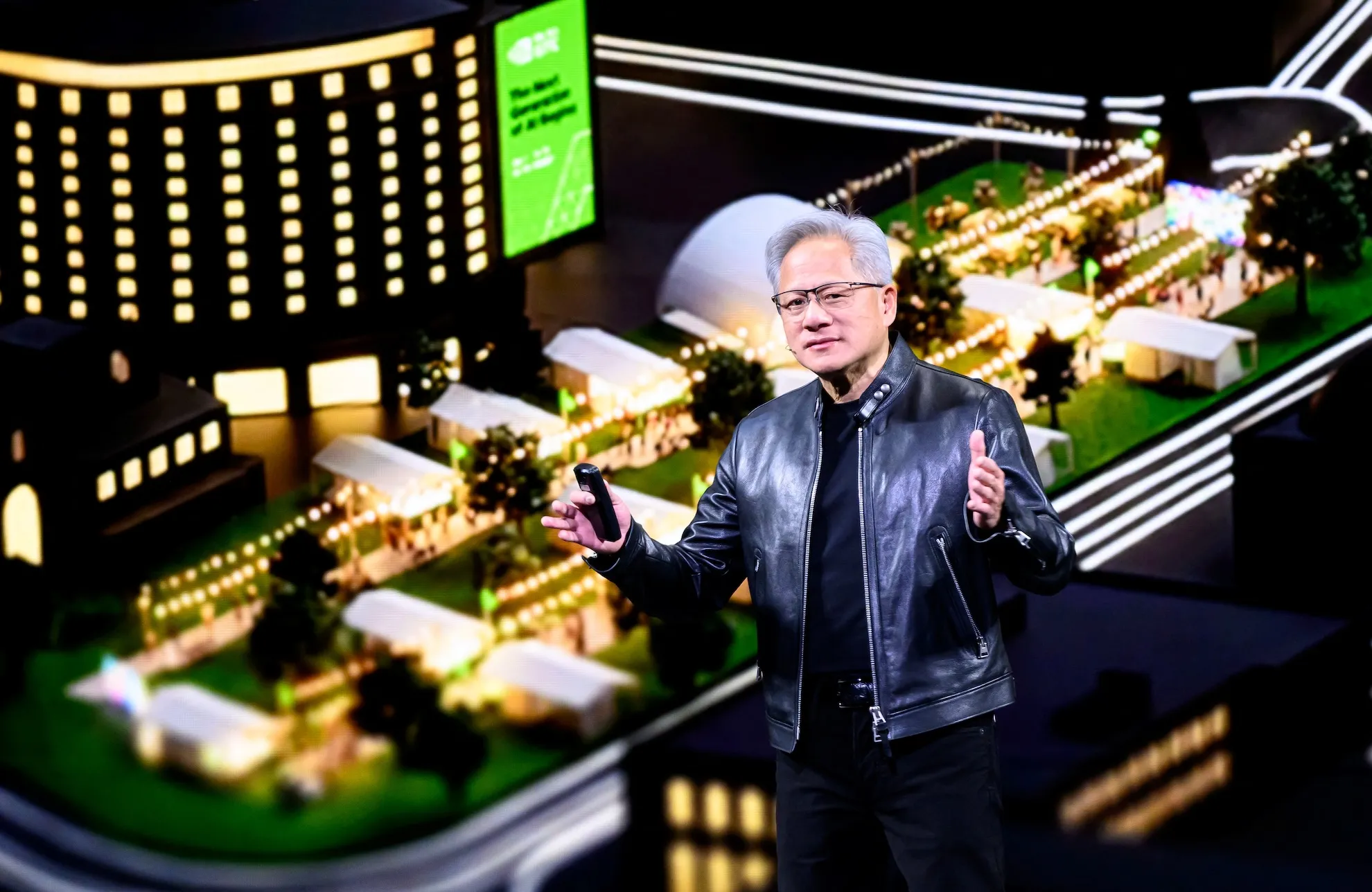 Man in black leather jacket speaking before a brightly lit technology event display