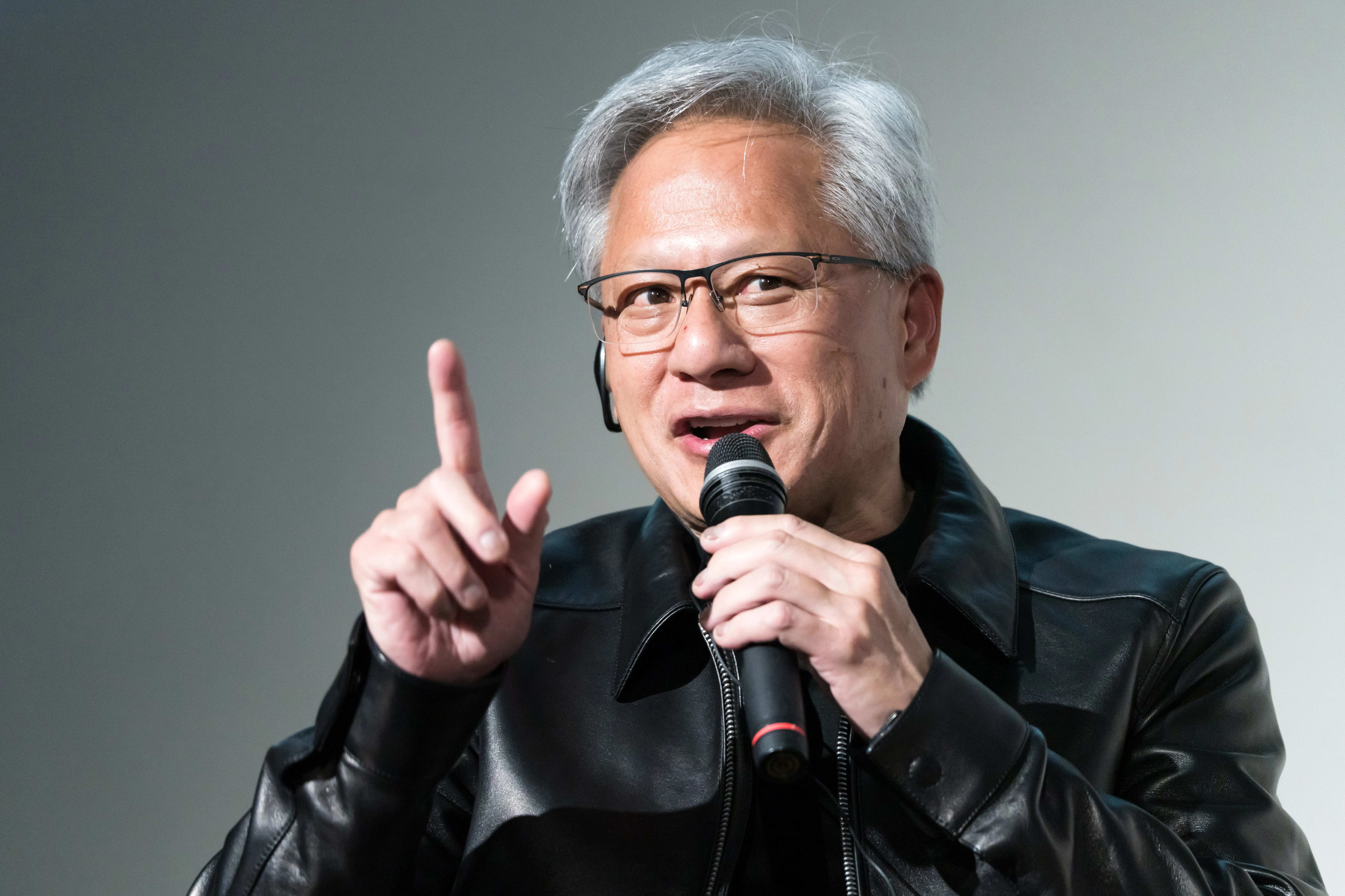 Man in glasses and black leather jacket speaks into a microphone, raising one finger.