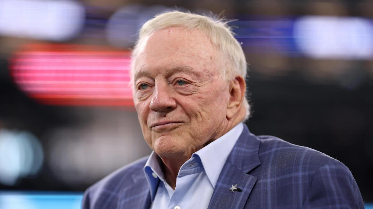 Older white-haired man in a blue checked blazer and pale shirt, standing against a blurred stadium backdrop.