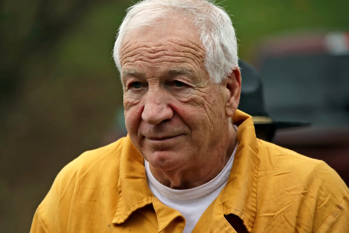 Older white-haired man in a yellow jacket looks toward the camera outdoors.
