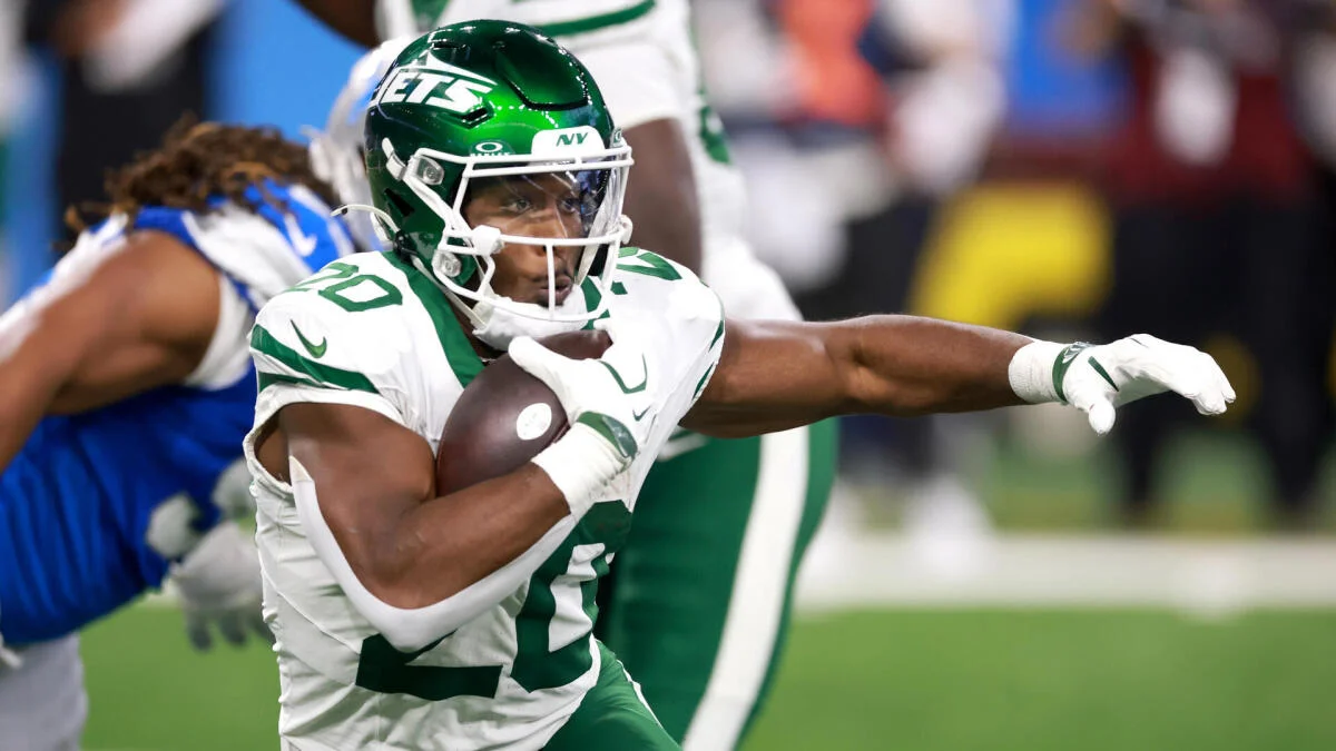 A Jets player in a green helmet and white uniform runs with the football as a Lions player pursues.