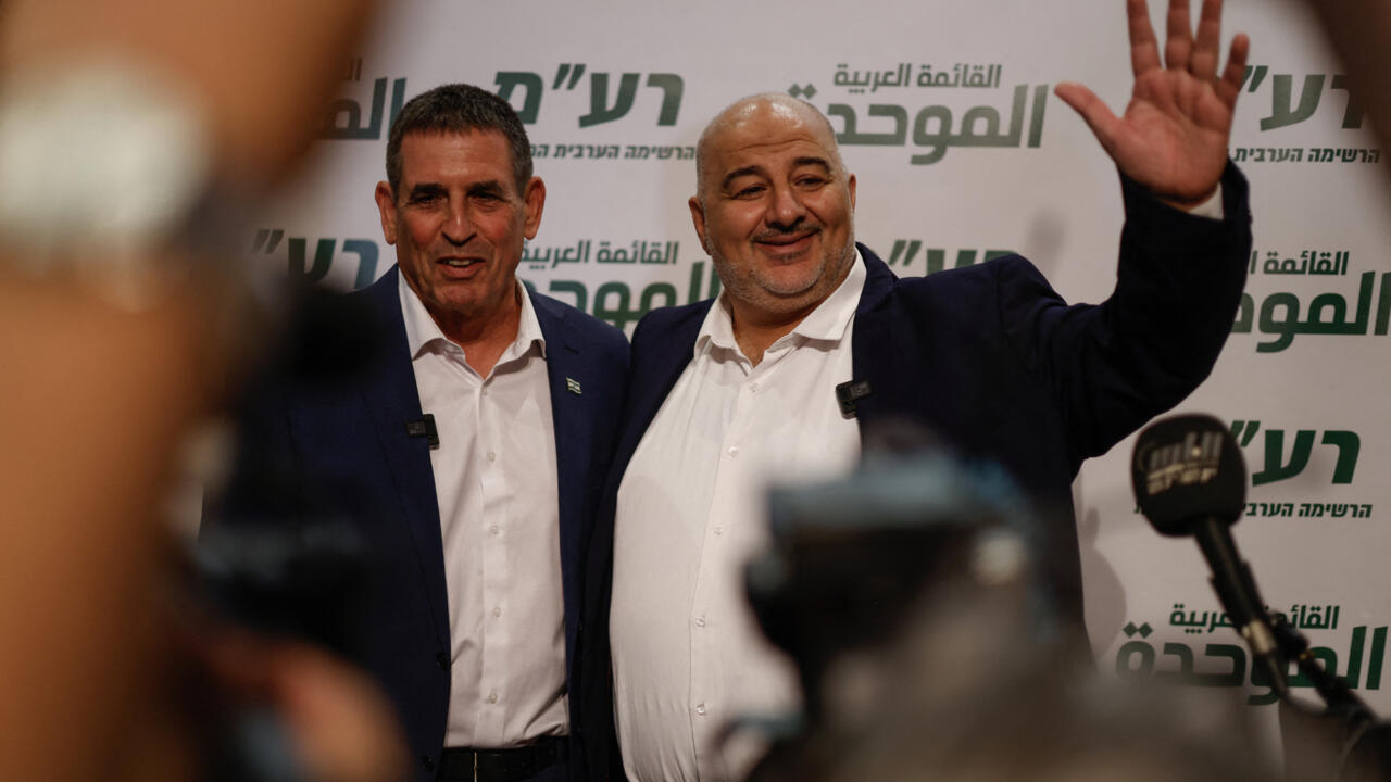 Two men in suits stand before Hebrew and Arabic campaign signage as one waves.