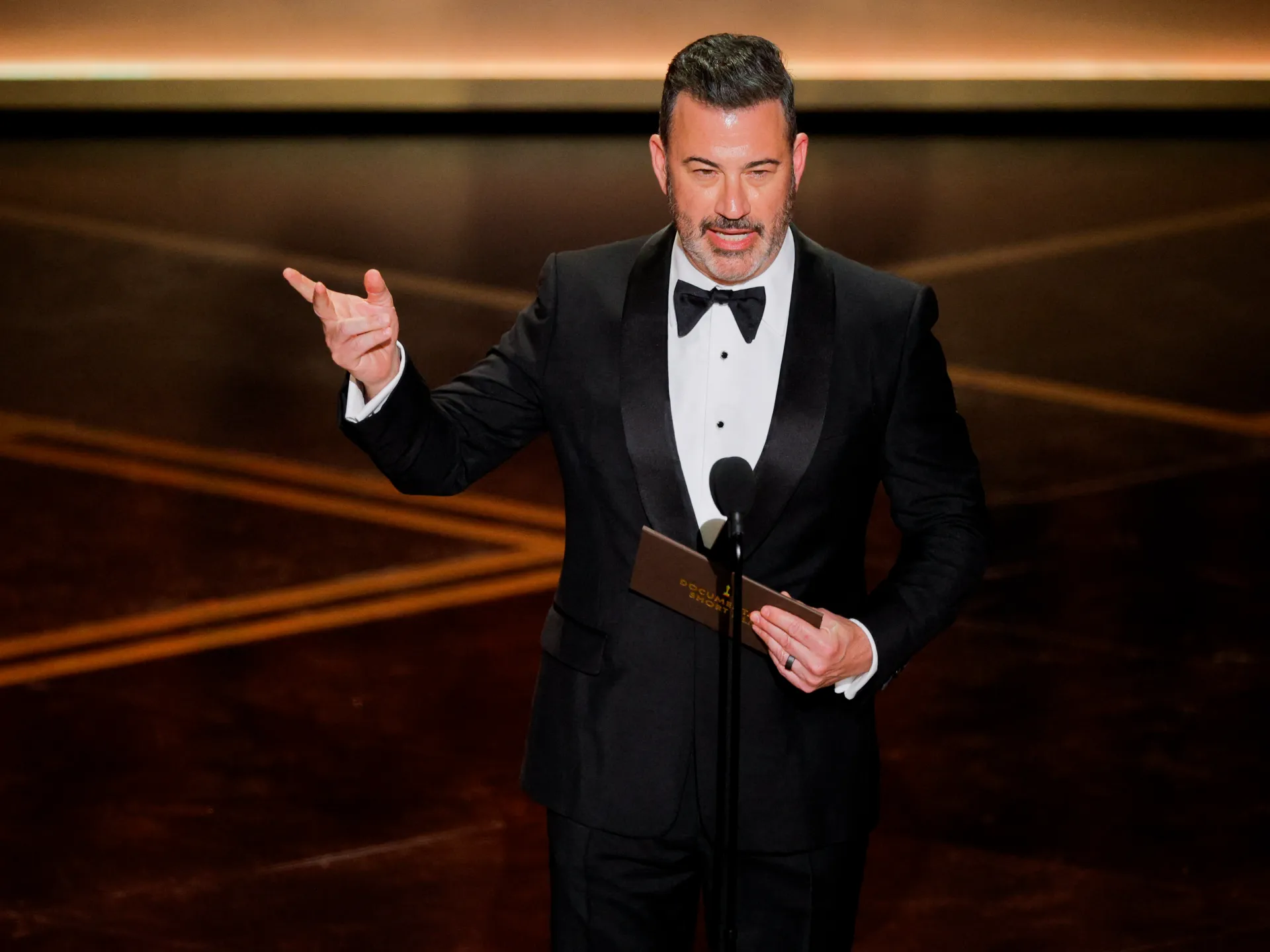 Man in tuxedo holding cards and speaking into a microphone on a dimly lit stage