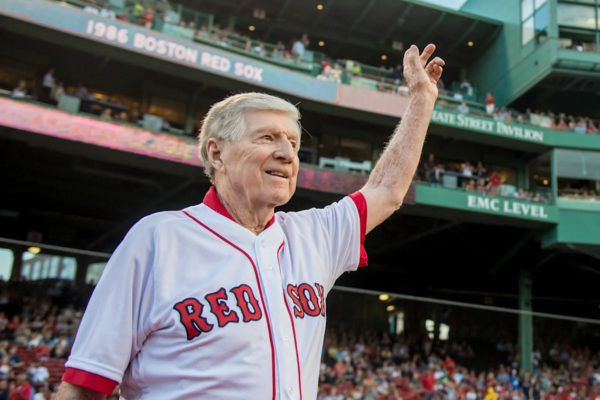 Elderly man in a Red Sox jersey waves from a baseball stadium field