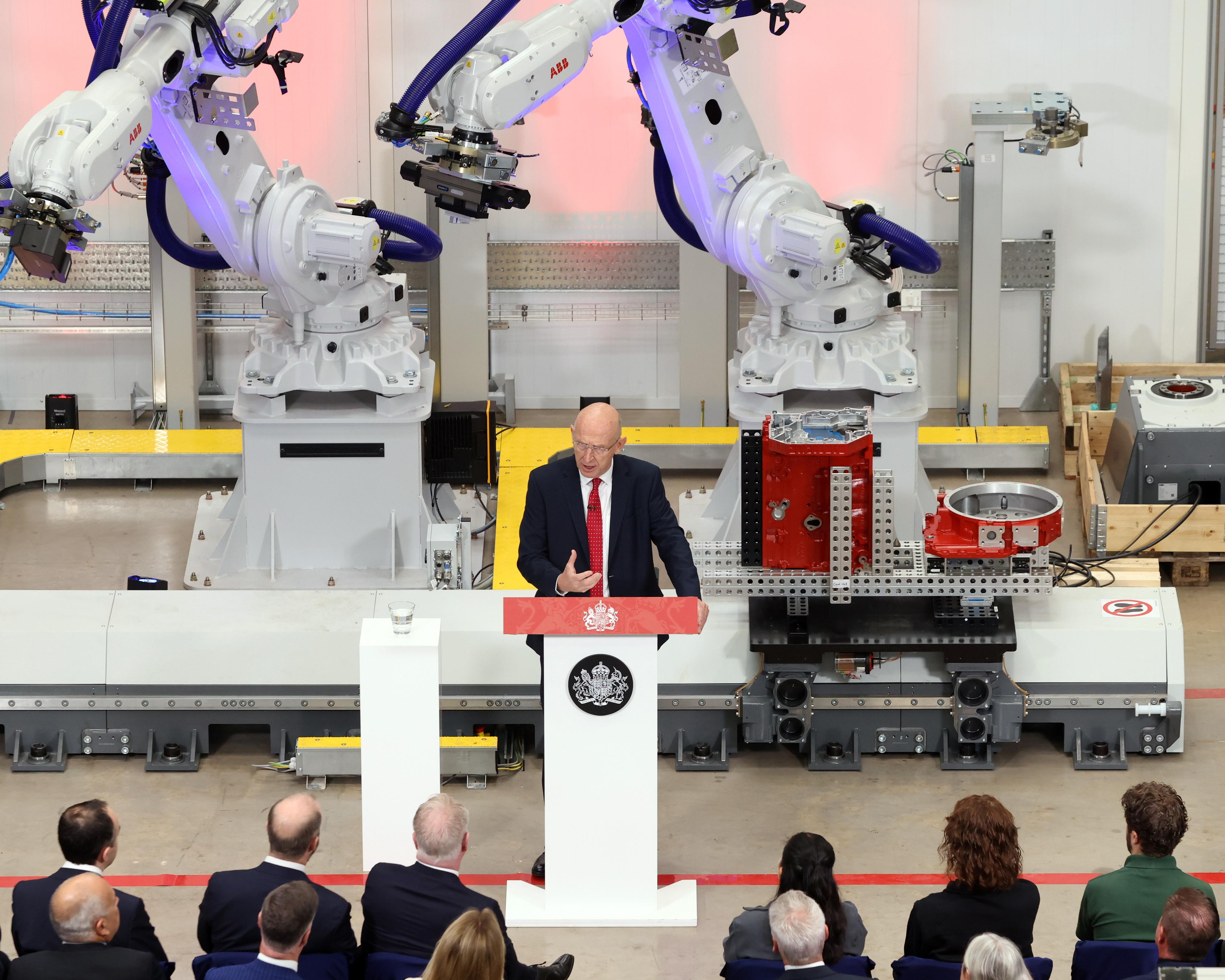 Man speaking at a podium in a factory surrounded by large industrial robotic arms