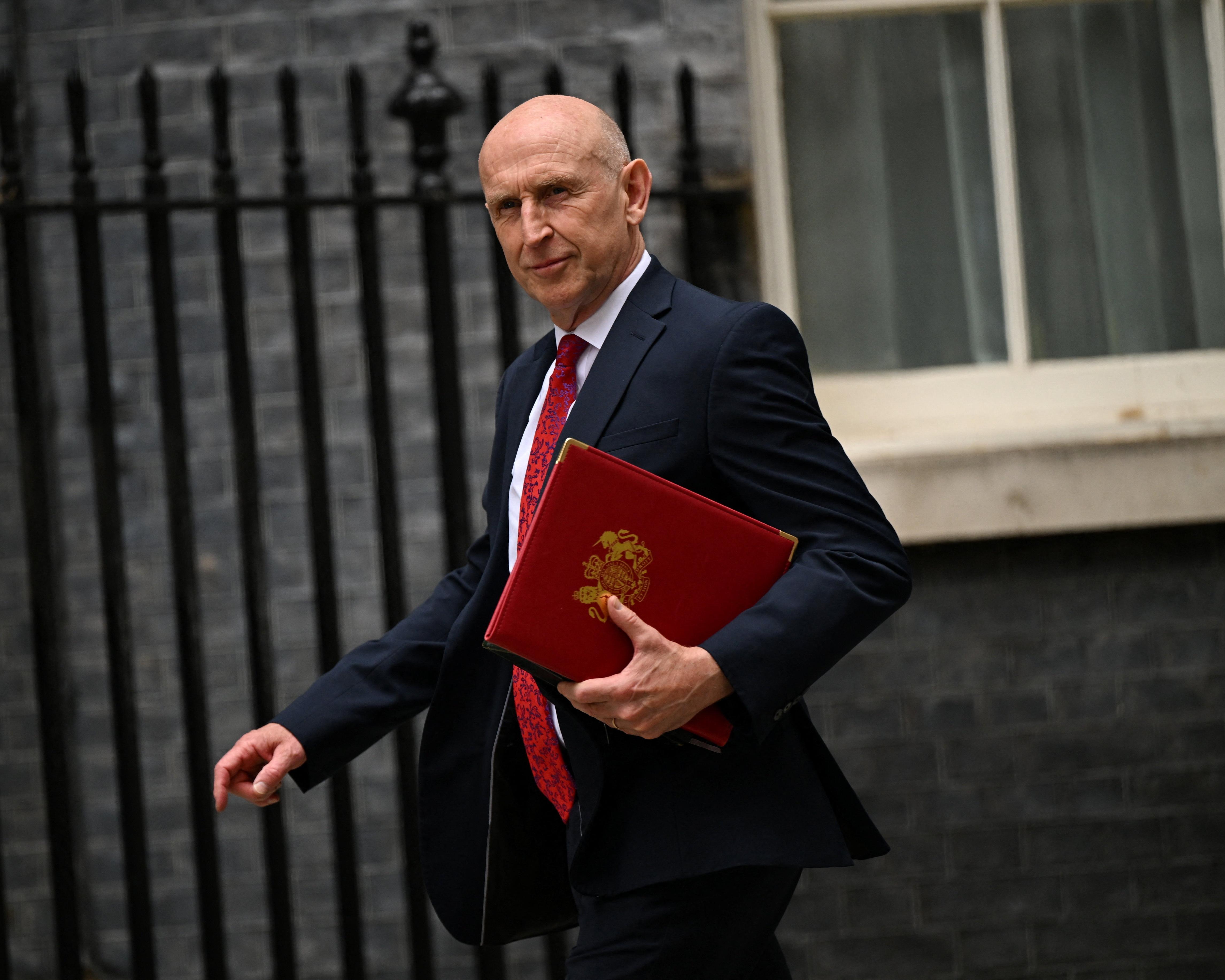 Bald man in dark suit carries a red official folder outside a building.