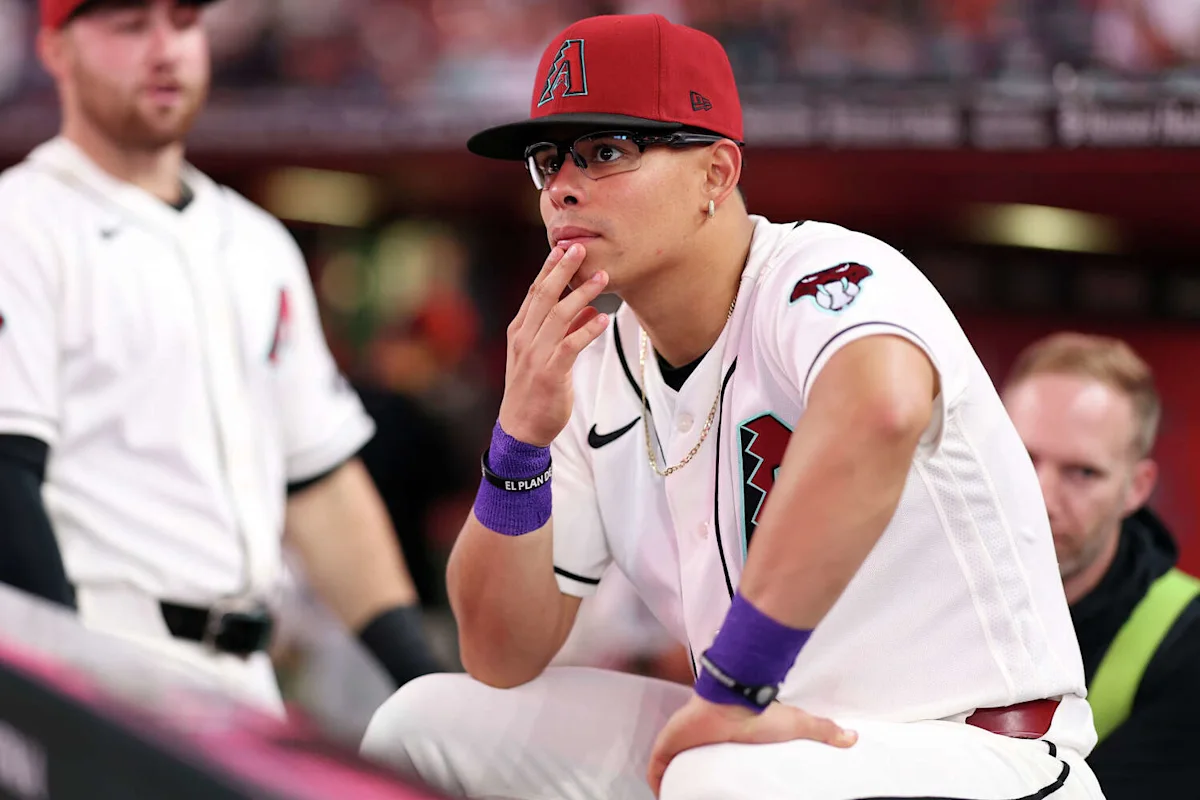 Baseball player in an Arizona Diamondbacks uniform sits in the dugout wearing sunglasses and a red cap.