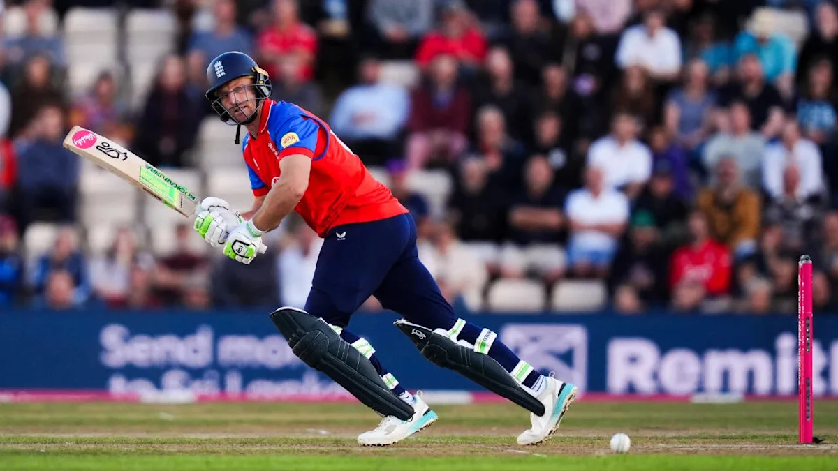 Cricketer in red and blue kit batting during a match as fielders’ stumps stand nearby