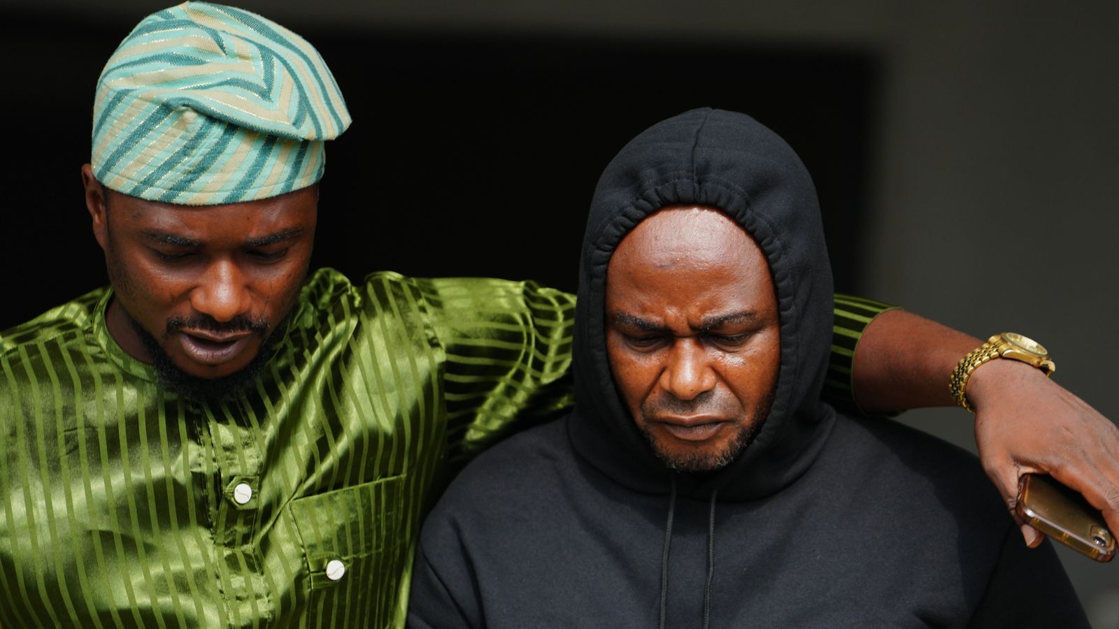 Two men sit closely together, both looking downward, with one wearing green traditional clothing and the other a black hoodie.