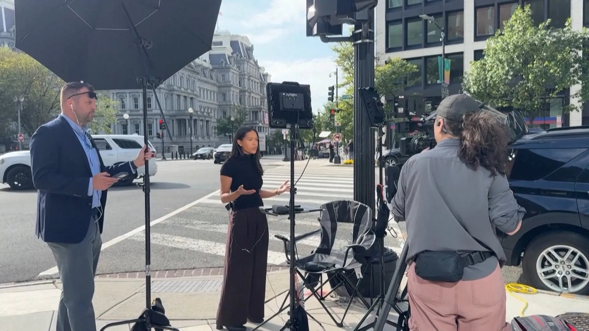 Camera crew films a woman speaking on a city sidewalk beside lighting equipment and a parked SUV.