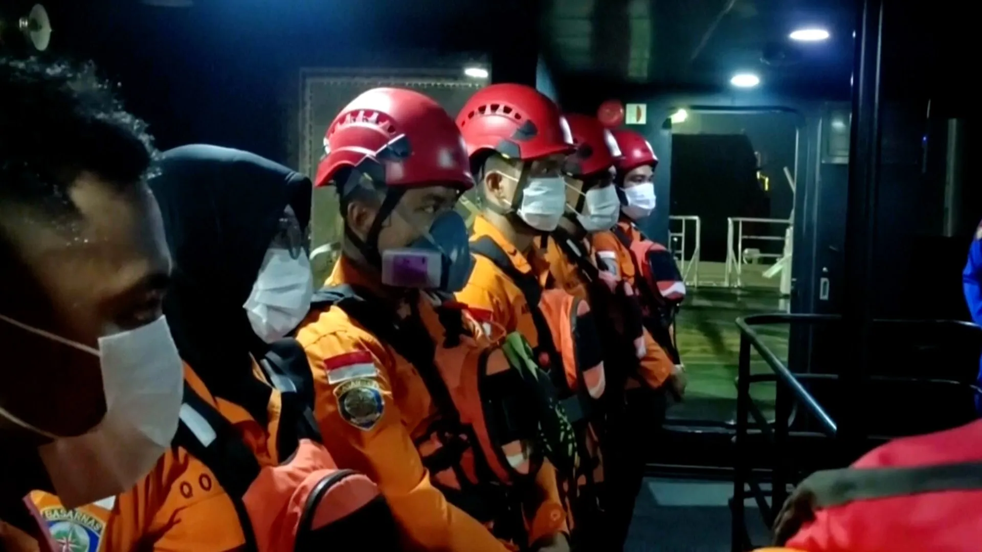 Masked emergency responders in orange uniforms and red helmets stand inside a dimly lit vehicle.
