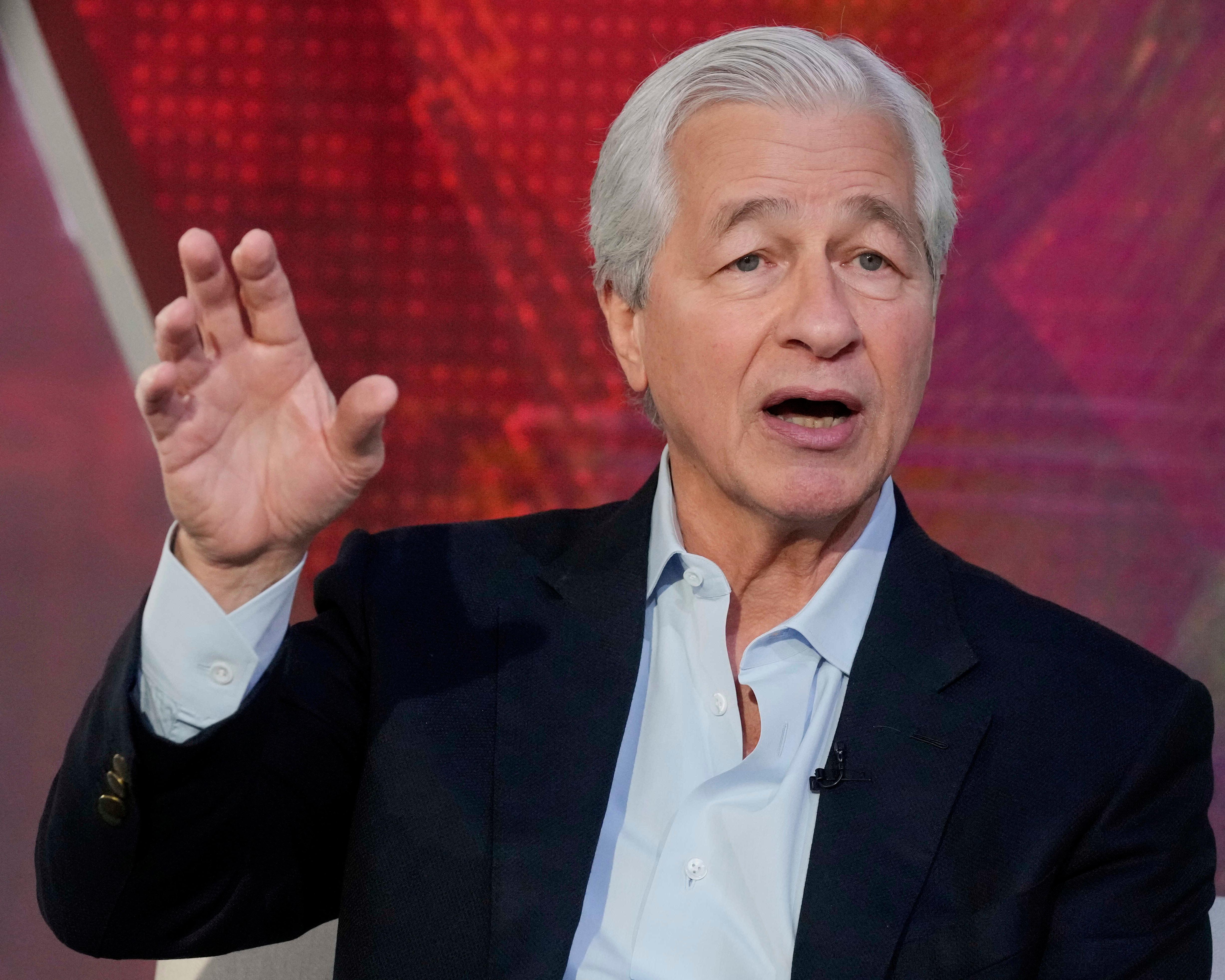 Silver-haired man in dark suit gestures with raised hand against a red abstract background