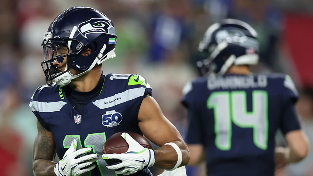 Seattle Seahawks player carries a football as a teammate stands blurred in the background.
