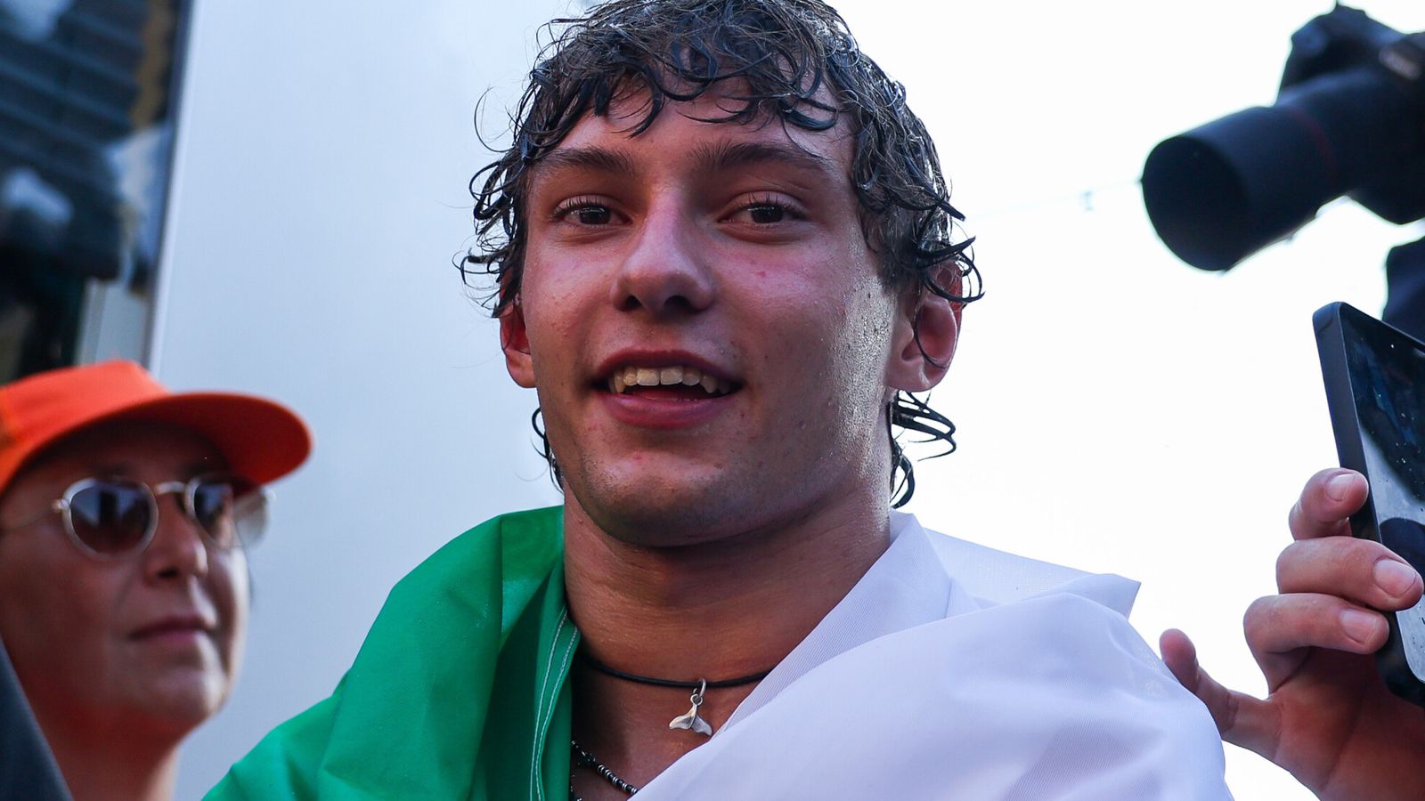 Smiling young man with wet hair wearing white clothing and a green garment amid cameras