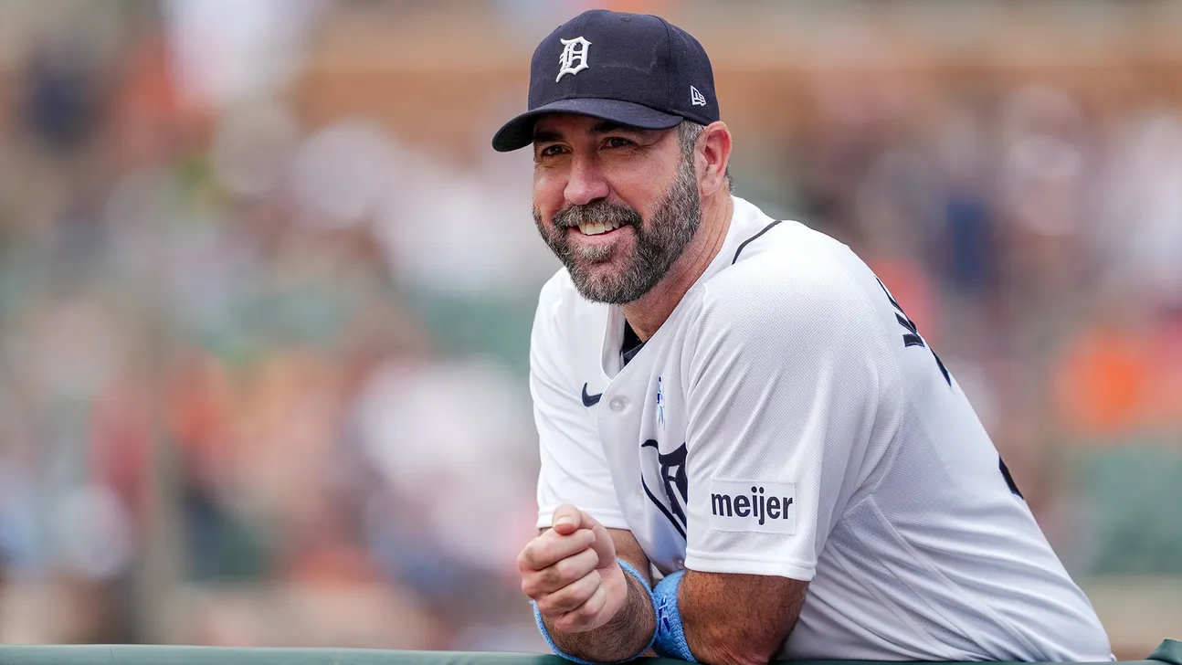 A bearded man in a Detroit Tigers cap and white baseball jersey leans forward against a blurred background.