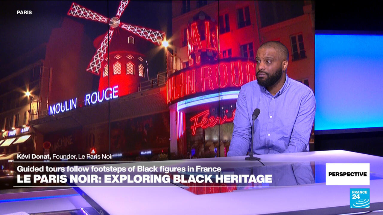 A bearded man in a blue shirt sits at a studio desk before a Paris-themed backdrop.
