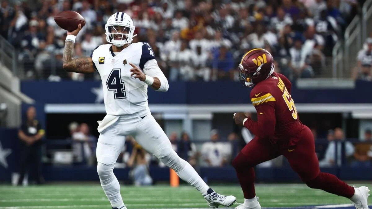 A quarterback in a white uniform prepares to throw as a burgundy-clad defender closes in.