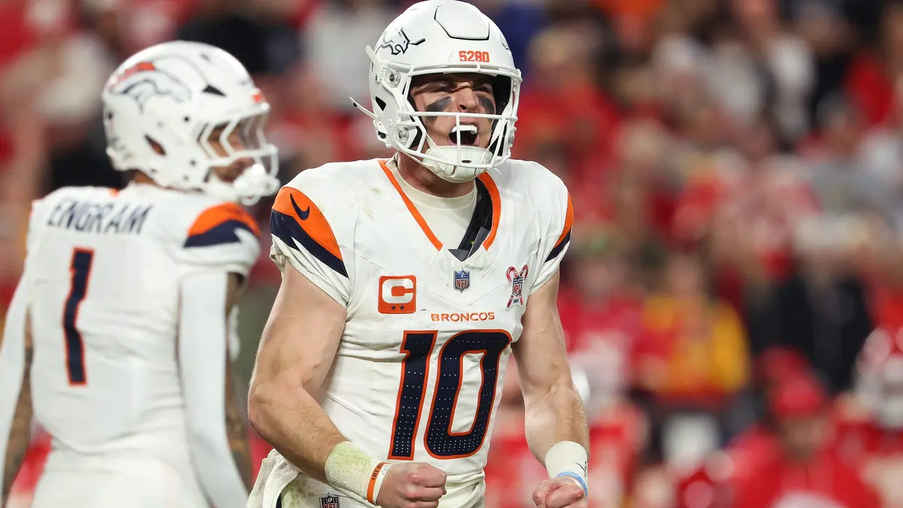 Denver Broncos player in white uniform shouts during a game as a teammate and crowd stand behind him.