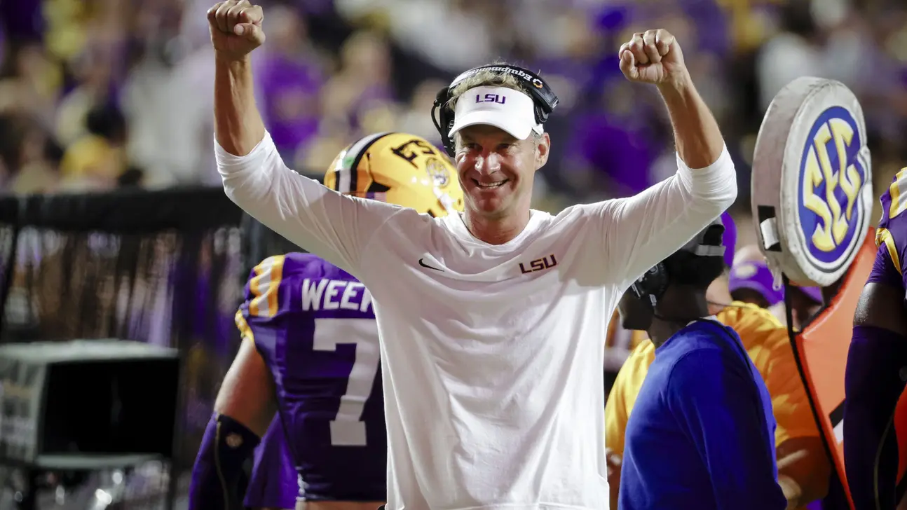 Football coach in white LSU shirt raises both fists amid players and fans